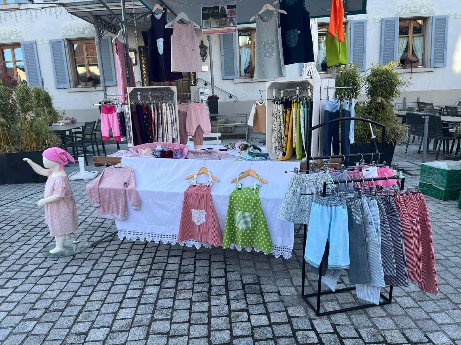 Marktstand von Suech dä Stern mit Kinderkleidern auf Bügeln und einer Kleiderpuppe